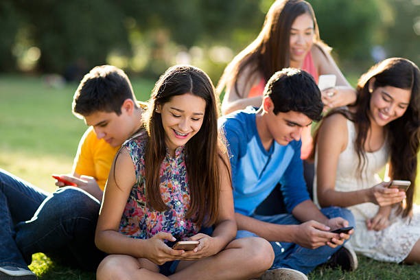 A group of young friends sitting together on the grass, chatting and looking at their phones